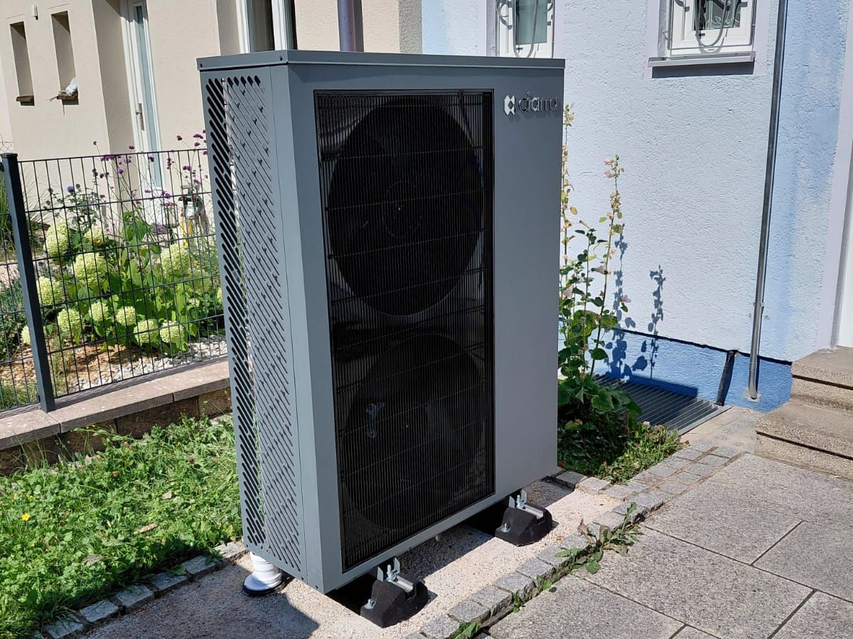 Graue Ciarra-Außeneinheit seitlich an der hellblauen Hauswand, daneben ein Vorgarten mit Zaun
